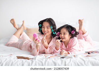 Happy young japanese woman and teenage girl in pajamas and curlers lie with cosmetics on bed, have fun, doing makeup in bedroom interior. Mother and daughter testing cosmetics at beauty day at home