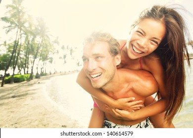 Happy Young Joyful Couple Having Beach Fun Piggybacking Laughing Together During Summer Holidays Vacation On Tropical Beach. Beautiful Energetic Fresh Interracial Multi-ethnic Couple, Man And Woman.