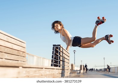 Happy Young Joyful Woman Doing A Balancing Act Wearing Rollerskates. Young Woman In Skates Jumping With The Support Of A Bench. Beautiful Young Happy Woman Wearing Roller Blades Doing A Stunt. 
