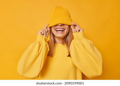 Happy young joyful woman hides eyes with hat smiles broadly shows white teeth foolishes around dressed in casual loose jumper isolated over yellow background. People and sincere emotions concept - Shutterstock ID 2090199001