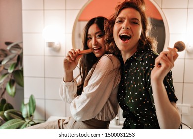 Happy Young Ladies Having Fun While Doing Makeup In Bathroom. Indoor Shot Of Multicultural Friends Singing In Front Of Mirror.