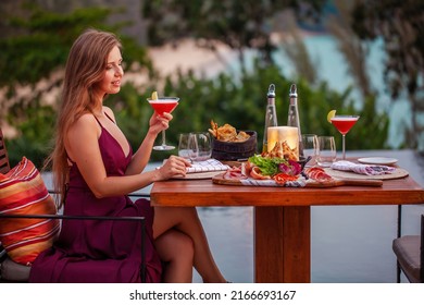 Happy Young Lady With A Glass Of Cocktail In Fine Dining Outdoor Restaurant. Sexy Girl In Elegant Evening Dress Celebrating Happy Valentines Day Holiday. Romantic Dinner On Sunset