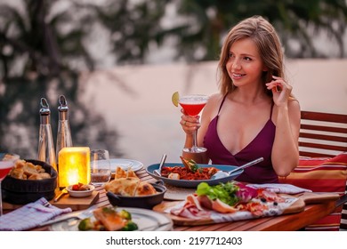 Happy Young Lady With A Glass Of Cocktail In Fine Dining Outdoor Restaurant. Sexy Girl In Elegant Evening Dress Celebrating Happy Valentines Day Holiday. Romantic Dinner On Sunset