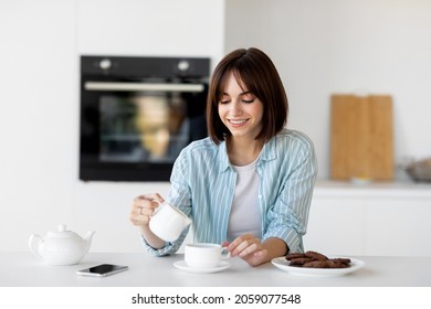 Happy young lady pouring milk in cup of coffee, sitting in kitchen interior, enjoying morning hot beverage and start of new day, empty space