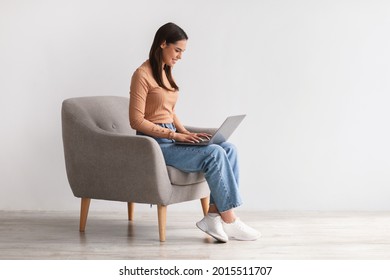 Happy young lady sitting in armchair, using laptop, studying remotely or having business meeting, free space. Millennial Caucasian woman having online class or watching webinar