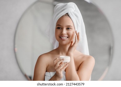 Happy Young Lady Using Face Cream, Looking At Mirror And Smiling, Holding Cream Jar And Applying Beauty Product On Her Cheeks After Morning Shower