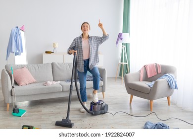 Happy Young Lady With Vacuum Cleaner Having Cleanup Idea At Messy Room After Party. Millennial Female Student Hoovering Dirty Apartment, Pointing Finger Up, Gesturing Eureka