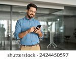 Happy young latin business man holding smartphone standing in office. Smiling hispanic businessman entrepreneur or manager using financial banking apps on cell phone technology device at work.