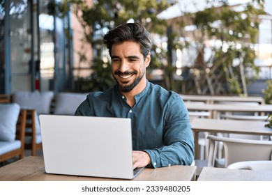 Happy young Latin business man using laptop sitting outdoors. Smiling Hispanic guy student or professional looking at computer sitting in city cafe elearning or hybrid working, searching job online. - Powered by Shutterstock