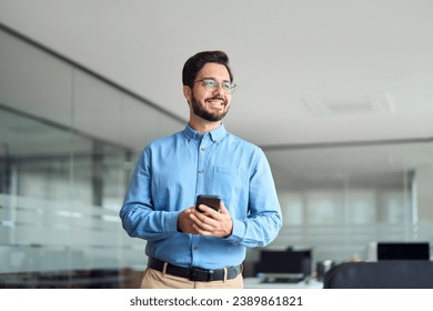 Happy young latin business man holding smartphone standing in office. Smiling hispanic businessman entrepreneur or manager using finances management app on cell phone technology looking away at work. - Powered by Shutterstock