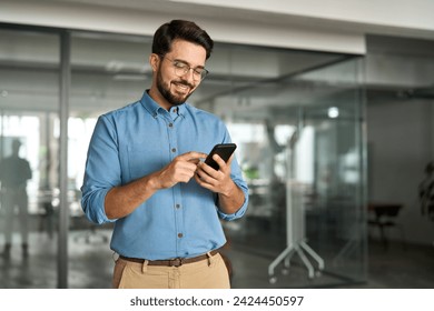 Happy young latin business man holding smartphone standing in office. Smiling hispanic businessman entrepreneur or manager using financial banking apps on cell phone technology device at work.