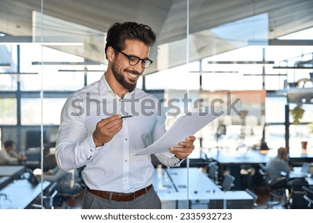 Happy young Latin business man checking financial documents in office. Smiling male professional account manager executive holding corporate tax bill papers reading plan overview standing at work.