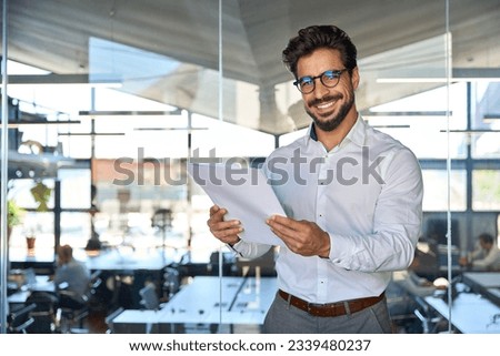 Happy young Latin business man checking financial documents in office. Smiling male professional account manager executive lawyer holding corporate tax bill papers standing at work. Portrait