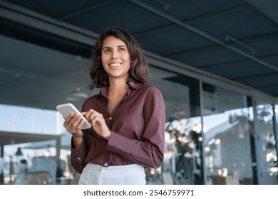 Happy young Latin Hispanic business woman holding mobile cell phone, watching app for work. Smiling middle eastern entrepreneur businesswoman holding smartphone cellphone standing in modern office - Powered by Shutterstock