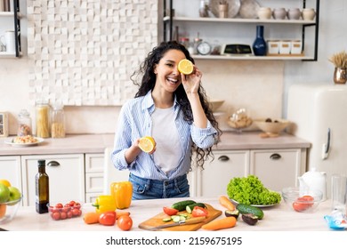 Happy Young Latin Lady Having Fun And Covering Eyes With Lemon Halves, Fooling Around In Kitchen Interior, Looking And Smiling At Camera While Cooking Dinner