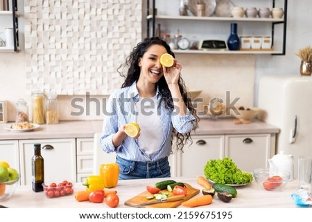 Happy young latin lady having fun and covering eyes with lemon halves, fooling around in kitchen interior, looking and smiling at camera while cooking dinner