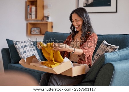Happy young latin woman checking clothes at home. Cheerful shopaholic hispanic woman checking clothes in package at home. Smiling mixed race girl unpacking parcel box with clothe after shopping online