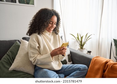 Happy young latin woman sitting on sofa holding mobile phone using cellphone technology doing ecommerce shopping, buying online, texting messages relaxing on couch in cozy living room at home. - Powered by Shutterstock