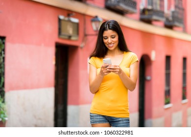 Happy young latin woman talking and texting on smart phone, wearing casual clothes sitting in with a take away coffee in a urban bench 