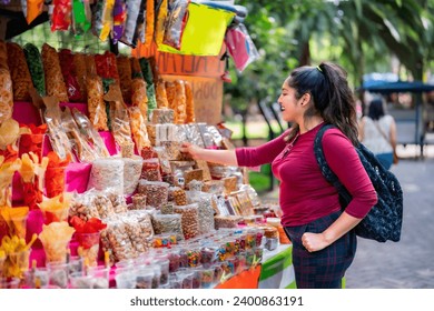 Happy young Latin woman tourist in casual clothes standing with backpack and holding traditional Mexican sweet candies while standing near stall on street market - Powered by Shutterstock