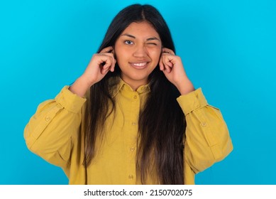 Happy Young Latin Woman Wearing Yellow Shirt Over Blue Background Ignores Loud Music And Plugs Ears With Fingers Asks To Turn Off Sound