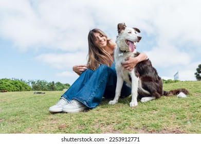 happy young latina colombian woman with long hair, dressed in blue jean pants is sitting on the grass in the park with her dog enjoying the weekend - Shutterstock ID 2253912771