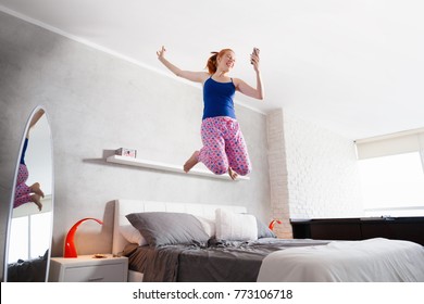 Happy Young Latina Woman Jumping Mid Air On Bed, Hispanic Girl Smiling And Laughing In Bedroom After Reading Good News On Mobile Phone.