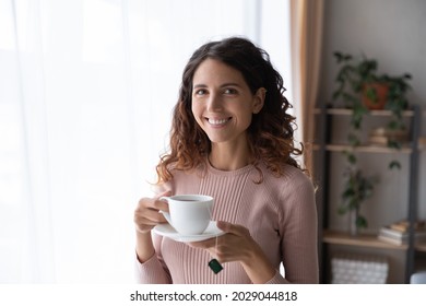 Happy Young Latina Woman Standing In Cozy Room Smile Looks At Camera Holds Cup Of Fresh Brewed Tea, Enjoy No Stress Calm Positive Pastime, New Day Drink Favourite Beverage. Leisure, Lifestyle Concept