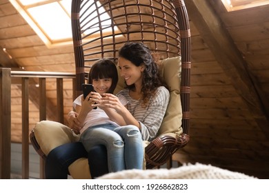 Happy Young Latino Mom And Small Daughter Relax In Cozy Chair Use Smartphone Gadget Together. Smiling Hispanic Mother And Little 8s Girl Child Sit In Rattan Furniture Have Fun Browse Cellphone.