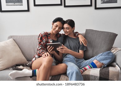 Happy young lesbian couple hugging having fun using digital tablet relaxing on couch at home. Two smiling women friends holding computer looking at screen enjoying surfing online watching videos. - Powered by Shutterstock
