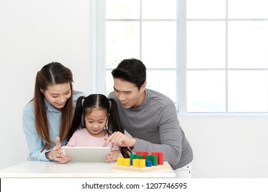 Happy young little asian cute girl watching or playing digital tablet, laptop or mobile with cheerful parents on desk in family time concept in white room with window. Preschool children learning. - Powered by Shutterstock