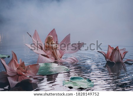 Happy young little blonde woman sits in huge pink flower lotus on water. Fantasy artistic photo. Beautiful girl in a fairy tale lake. Girl Thumbelina. Image Goddess of nature in blue mystic river fog