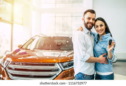 Happy young lovely couple in casual wear hugging while buying first new family car together in dealership. Woman shows car key on camera