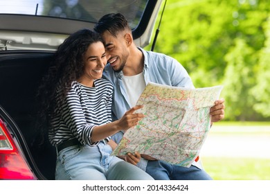 Happy young lovers cuddling while sitting on auto trunk and watching road map, copy space. Handsome arab guy bonding with his beautiful girlfriend, having break while car trip together
