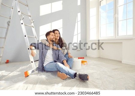 Happy young loving couple planning home repair while sitting on floor in living room. Cheerful wife hugging her husband in freshly painted room. Home repair and decoration concept