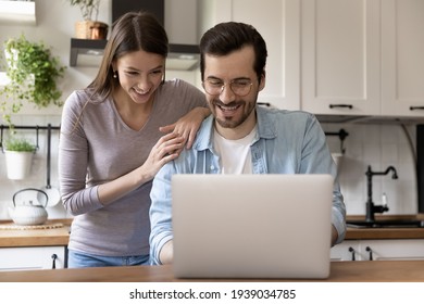 Happy Young Loving Family Couple Looking At Laptop Screen, Reading E-mail With Good News. Smiling Handsome Man Showing Funny Video On Computer To Pretty Joyful Wife, Sitting At Table In Kitchen.