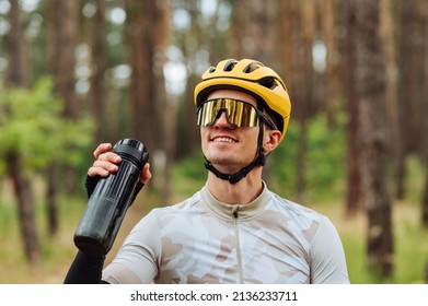 Happy young male cyclist in sportswear wearing helmet and goggles stands in forest with bottle of water in hands and smiling. - Powered by Shutterstock