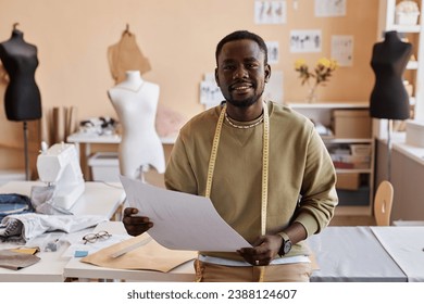 Happy young male fashion designer with sketch of new attire looking at camera while standing by workplace against group of mannequins - Powered by Shutterstock