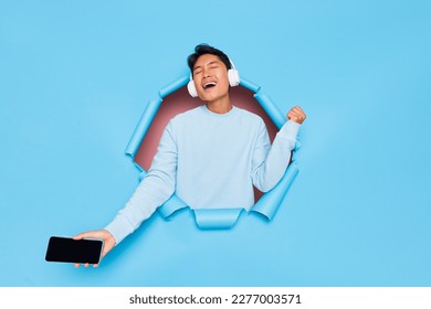 Happy young male model wearing white sweatshirt and headphones posing on blue studio background, student listening music, lifestyle concept, copy space.