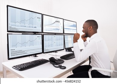 Happy Young Male Stock Market Broker Looking At Graphs On Multiple Computer In Office