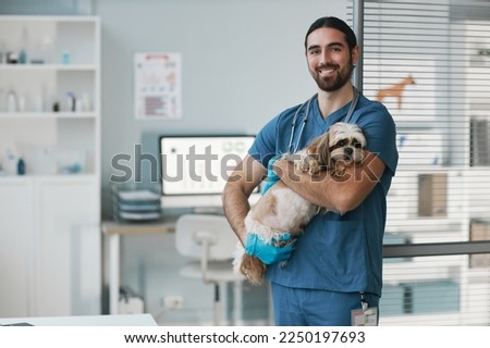 Happy young male veterinarian in blue uniform holding cute yorkshire terrier and looking at camera while standing in medical office