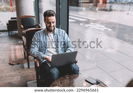 Happy young male wearing casual outfit sitting with netbook on laps and typing on keyboard while having break in contemporary cafeteria near glass wall