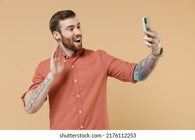 Happy young man 20s he wears orange shirt hold in hand use mobile cell phone meet greet communicate waving hand isolated on plain pastel light beige background studio portrait. Tattoo translate fun