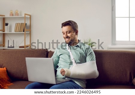 Happy young man with arm in a cast sitting on the couch at home and communicating on a laptop. Man in rehab after injury at home watching video or chatting on computer and smiling.