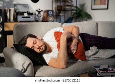 Happy young man with beard fell asleep on the couch in the living room. The sun shines on his face. The man hugged pillow with hands. The brunette is dressed in pajamas and lying on blanket.