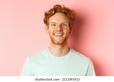 Happy young man with beard and messsy red haircut, smiling with white teeth and cheerful expression, standing over pink background
