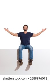 Happy Young Man In Boots, Jeans And Blue Shirt Is Sitting On A Top With Arms Outstretched, Looking Up And Smiling. Full Length Studio Shot Isolated On White.