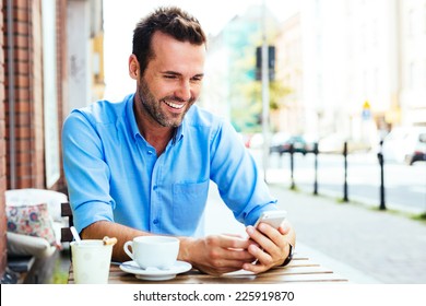 Happy Young Man Browsing Internet On Smartphone Sitting At Outdoor Cafe.