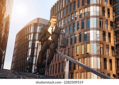Happy young man businessman in suit with briefcase having fun near business building corporate company, slides down railing of stairs. Joyful guy near office house in financial district. Copy ad space