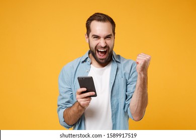 Happy young man in casual blue shirt posing isolated on yellow orange wall background studio portrait. People lifestyle concept. Mock up copy space. Hold mobile phone, doing winner gesture, screaming